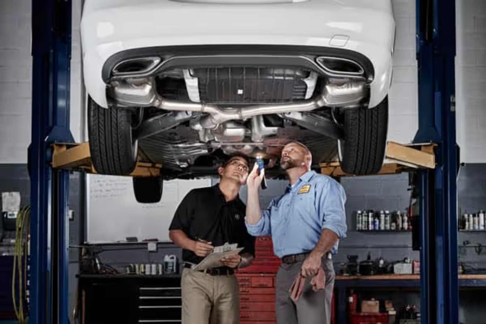 Two mechanics inspect the undercarriage of a white Mercedes-Benz on a lift.