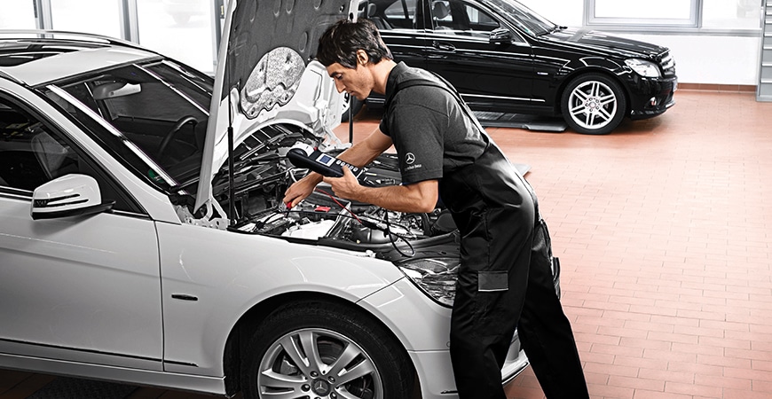 A mechanic in black overalls diagnoses a white Mercedes-Benz with a diagnostic tool.