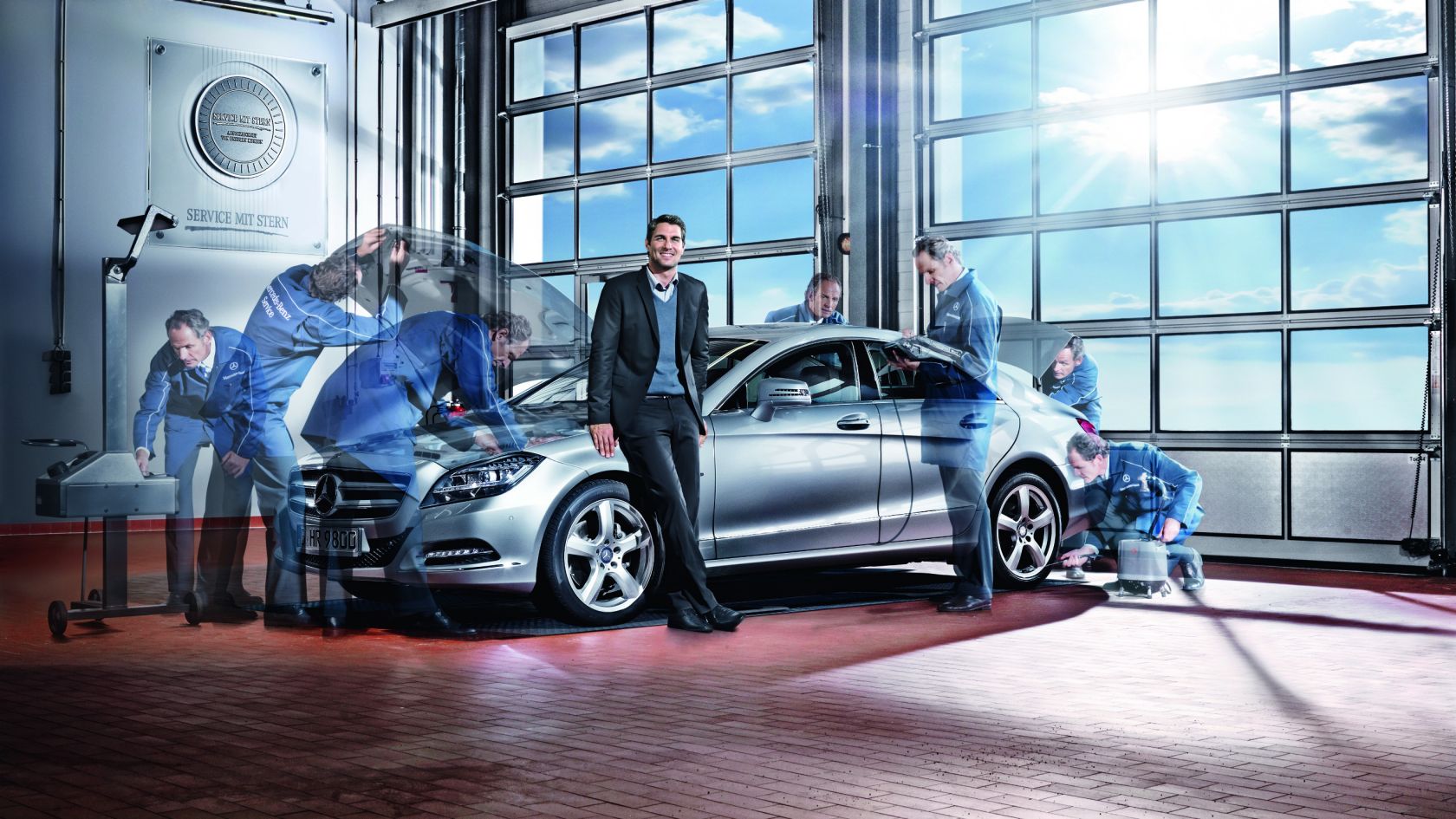 A businessman stands next to a silver Mercedes-Benz with mechanics working around it.