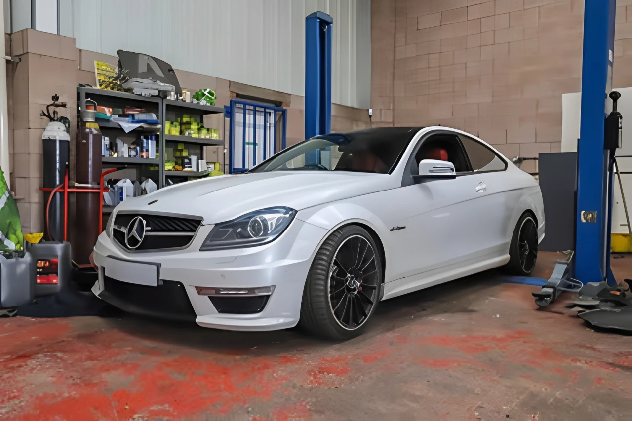 A white Mercedes-Benz C63 AMG coupe is parked inside an auto repair shop.